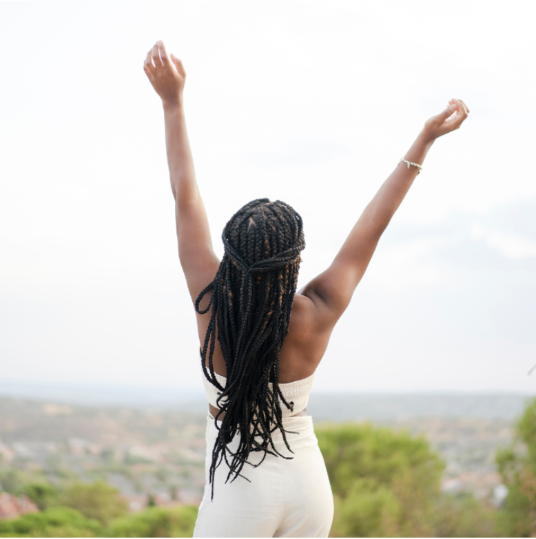 A woman with long braided hair stands outdoors with arms raised in triumph, overlooking a scenic landscape—representing how to heal from trauma and abuse through freedom, empowerment, and personal restoration.